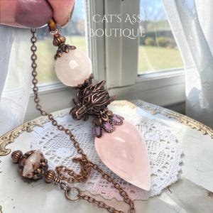 Puede incluir: Un péndulo de cuarzo rosa con cadena y detalles en cobre. El péndulo presenta un diseño de cabeza de dragón y un cristal en forma de lágrima. El texto "CAT'S ASS BOUTIQUE" es visible en el fondo.