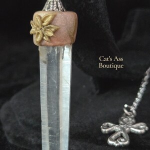 May include: A clear quartz crystal pendant with a silver-colored chain and a decorative cross charm. The pendant features a light brown top with a gold-colored flower accent. The text "Cat's Ass Boutique" is visible.