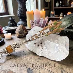 Puede incluir: Una varita decorativa con un mango plateado y una gema naranja en la base. La varita presenta un diseño verde y marrón y descansa sobre un gran cristal transparente. El texto "CAT'S ASS BOUTIQUE" es visible.