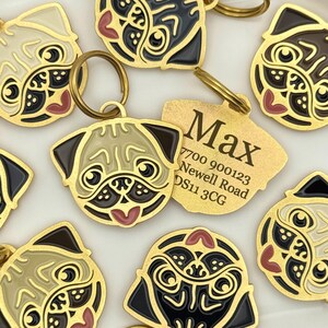 May include: A collection of gold-toned metal pet ID tags shaped like pug faces with black, cream, and pink enamel details. One tag is flipped over to reveal the engraved text: Max, 7700 900123, Newell Road, DS11 3CG. Each tag includes a circular ring.