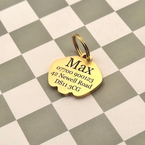 May include: A gold-colored pet tag in a unique shape, with a ring for attachment. The tag has the name "Max", a phone number, and an address engraved in black. The background is a checkered pattern of gray and white squares.