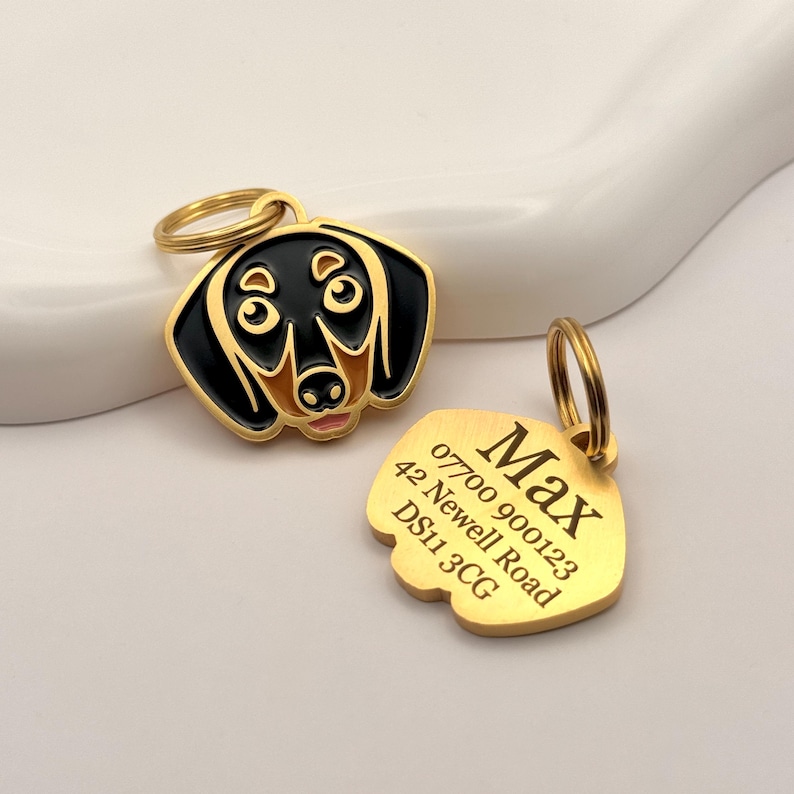 May include: Two gold-colored pet tags. One tag is shaped like a dachshund dog's head, with black and tan enamel. The other tag is gold-colored with the name "Max" and contact information engraved on it.