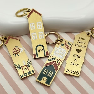 May include: A collection of gold-colored house-shaped keychains. Each keychain features unique designs, including house numbers, floral accents, and the text "Home Sweet Home," "Our New Home," and names. The keychains have gold key rings.