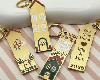 Könnte beinhalten: Eine Sammlung goldfarbener Schlüsselanhänger in Hausform. Jeder Schlüsselanhänger hat einzigartige Designs, darunter Hausnummern, florale Akzente und die Texte "Home Sweet Home", "Our New Home" und Namen. Die Schlüsselanhänger haben goldene Schlüsselringe.