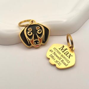 May include: Two gold-colored dog tags. One tag is shaped like a dachshund's head, with black, brown, and gold enamel. The other tag is gold with the name "Max" and an address engraved on it.
