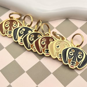 May include: A collection of dachshund-shaped keychains in various colors, including brown, black, and tan. Each keychain has a gold-colored ring and a detailed face design. The keychains are arranged on a checkered surface.