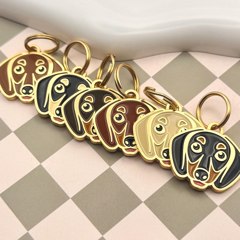 May include: A collection of dachshund-shaped keychains in various colors, including brown, black, and tan. Each keychain has a gold-colored ring and a detailed face design. The keychains are arranged on a checkered surface.