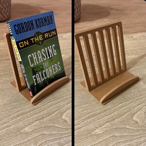 May include: A wooden book stand in a light brown color, holding a book titled "ON THE RUN CHASING THE FALCONERS" by Gordon Korman. The stand has a curved base and vertical slats. The book's cover is blue, green, and white.