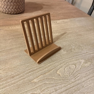 Puede incluir: Un atril de madera marrón claro con respaldo de listones y base curva. El soporte está diseñado para sujetar libros o tabletas. El soporte está sobre una mesa de vetas de madera clara.