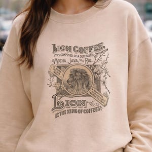 Op de afbeelding: Beige sweatshirt met een vintage Lion Coffee-ontwerp. Het ontwerp bevat een leeuwenkop, bloemmotieven en de tekst "Lion Coffee" en "The King of Coffees!"