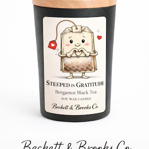 以下が含まれることがあります： 木製の蓋が付いた黒い大豆ワックスキャンドル。ラベルには、ハート型のタグが付いた漫画のティーバッグのキャラクターが描かれています。ラベルのテキストは「Steeped in Gratitude」と「Bergamot Black Tea」です。「Beckett & Brooks Co.」のブランド名も表示されています。