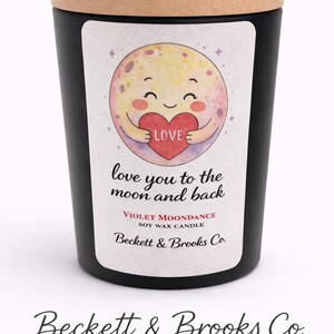 Op de afbeelding: Zwarte sojawas kaars met een houten deksel. Het etiket toont een lachende maan die een hart vasthoudt met het woord "LOVE". Tekst op het etiket luidt "love you to the moon and back" en "Violet Moondance Soy Wax Candle".