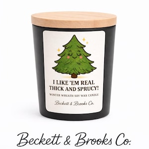 Könnte beinhalten: Schwarze Kerze mit Holzdeckel. Das Etikett zeigt eine lächelnde grüne Weihnachtsbaum-Illustration und den Text "I LIKE 'EM REAL THICK AND SPRUCY!" und "Winter Wreath Soy Wax Candle". Der Markenname "Beckett & Brooks Co." wird ebenfalls angezeigt.