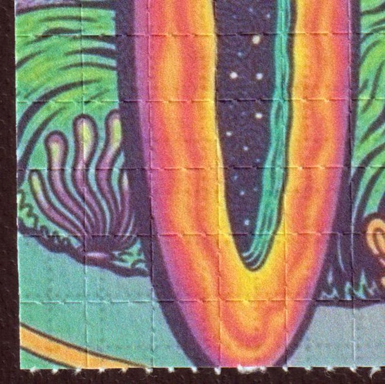 May include: Close-up of a colorful, psychedelic art print. The design features a vibrant gradient of orange, yellow, and purple hues, with a central, elongated shape. The artwork is divided into small squares, suggesting a perforated sheet.