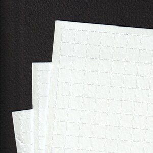 May include: Close-up of three sheets of white paper with a grid pattern. The paper has a textured surface and is stacked against a dark background. The grid pattern is made up of small, dashed lines.