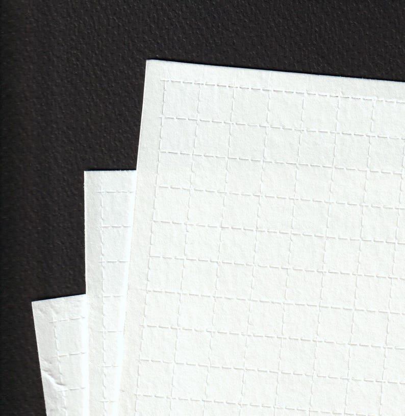 May include: Close-up of three sheets of white paper with a grid pattern. The paper has a textured surface and is stacked against a dark background. The grid pattern is made up of small, dashed lines.