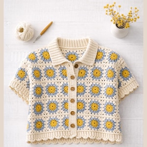 Può includere: Top all'uncinetto color crema con un motivo a margherita giallo e azzurro. La camicia a maniche corte ha un colletto e si chiude con bottoni in legno. Sono visibili un gomitolo di lana e un uncinetto.