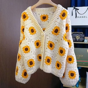 Op de afbeelding: Crèmekleurig gehaakt vest met een zonnebloemmotief. Het vest heeft een V-hals en lange mouwen. De zonnebloemen zijn geel met bruine centra. Het kledingstuk wordt op een houten hanger getoond.