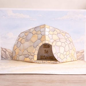 Puede incluir: Una tarjeta emergente de Pascua con una tumba de piedra con las palabras "Él ha resucitado" en su interior. La tarjeta está abierta, revelando una escena tridimensional con un cielo azul. La tarjeta está sobre una superficie de madera.