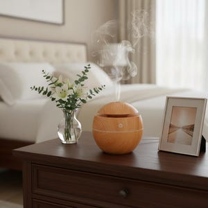 Op de afbeelding: Een houten diffuser voor etherische olie met een licht houtnerfpatroon staat op een donkerbruin nachtkastje. Een kleine glazen vaas met witte bloemen en een ingelijste foto staan ook op de tafel. Dampwolkjes stijgen op uit de diffuser.