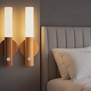 May include: Two wall-mounted sconces with a cylindrical white light fixture atop a wooden base. The sconces are mounted on a gray wall, with a bed and pillows in the background. Each sconce has a small sensor.