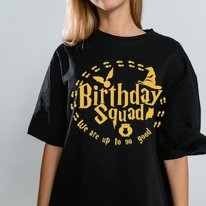 May include: Black t-shirt with a yellow graphic that says "Birthday Squad" with a wizard hat, a flying bird, and the words "We are up to no good". The shirt has short sleeves.