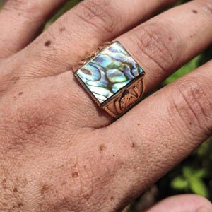 Könnte beinhalten: Ein goldfarbener Ring mit einer quadratischen Einlage aus irisierendem Abalone-Muschelmaterial. Die Oberfläche der Muschel zeigt wirbelnde Muster in Blau-, Grün- und Perlmutttönen. Der Metallring weist gravierte Details an den Seiten auf.