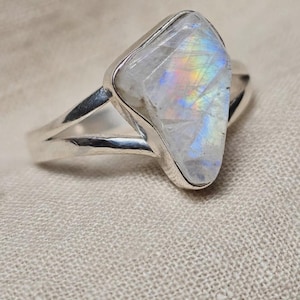 May include: A silver ring featuring a triangular, translucent moonstone with a visible iridescent blue and rainbow sheen. The stone is set in a smooth metal bezel, and the band splits into two separate silver arms that connect to the setting.