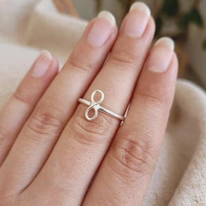 May include: A polished silver ring featuring a delicate infinity symbol centred on a thin, smooth band. The metallic surface reflects light, highlighting the continuous loop design of the infinity motif which sits horizontally across the finger.