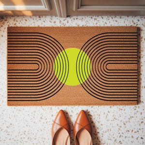 May include: A rectangular doormat with a geometric design. The mat features a brown background with a central neon green circle and black curved lines. Brown leather shoes are at the bottom of the image.