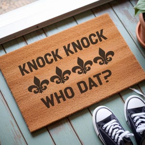 May include: A rectangular doormat made of natural brown coir fiber. The mat features the text "KNOCK KNOCK WHO DAT?" in dark brown, along with fleur-de-lis symbols. A pair of navy blue canvas sneakers with white soles and laces are visible.