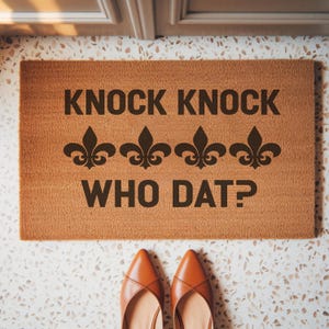 May include: A rectangular doormat with the text "KNOCK KNOCK WHO DAT?" in dark brown. Three fleur-de-lis symbols are between the phrases. The mat is a light brown color and is on a speckled white and gray floor. A pair of brown shoes is at the bottom.