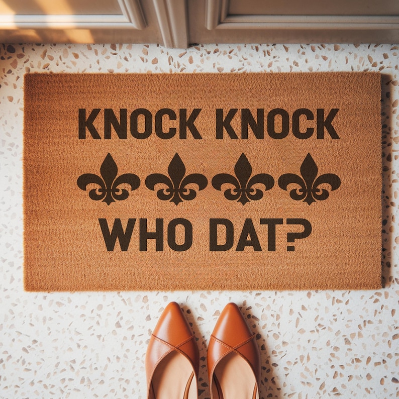 May include: A rectangular doormat with the text "KNOCK KNOCK WHO DAT?" in dark brown. Three fleur-de-lis symbols are between the phrases. The mat is a light brown color and is on a speckled white and gray floor. A pair of brown shoes is at the bottom.