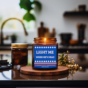 May include: A lit, brown glass jar candle with a blue label that reads "LIGHT ME WHEN HE'S DEAD" with white text and star accents. The candle sits on a wooden coaster, with a smaller jar and greenery in the background.