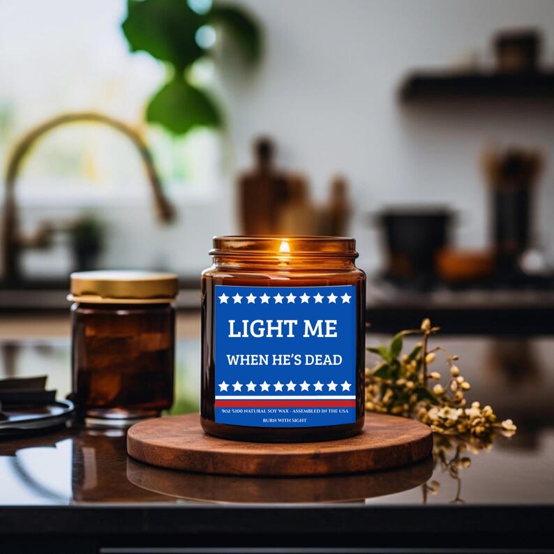 May include: A lit, brown glass jar candle with a blue label that reads "LIGHT ME WHEN HE'S DEAD" with white text and star accents. The candle sits on a wooden coaster, with a smaller jar and greenery in the background.