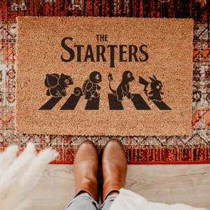 May include: A rectangular tan coir doormat featuring the black text THE STARTERS above a black graphic of four creatures walking across a crosswalk. The design includes silhouettes of Bulbasaur, Squirtle, Charmander, and Pikachu in a line.