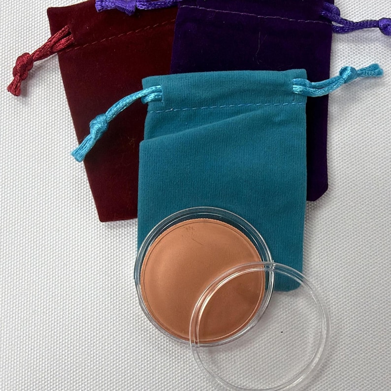 May include: An overhead shot shows three velvet drawstring pouches in burgundy, teal, and purple. A clear plastic container with a copper-colored disc inside is in front of the teal pouch. The pouches have matching colored drawstrings.