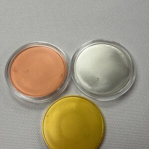 May include: Three round objects: one copper-colored, one silver-colored, and one gold-colored. Each is in a clear plastic container. The gold object is a solid disc. The background is a light gray fabric.