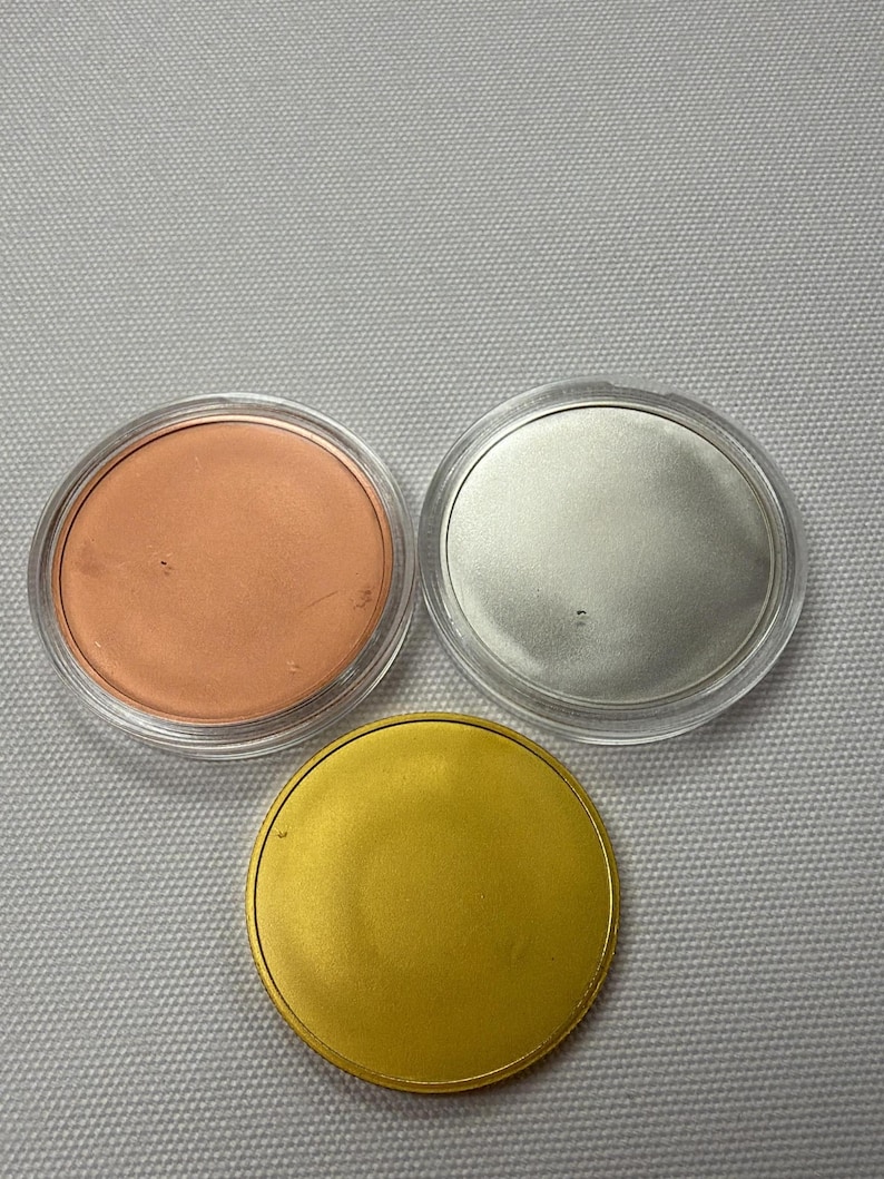 May include: Three round objects: one copper-colored, one silver-colored, and one gold-colored. Each is in a clear plastic container. The gold object is a solid disc. The background is a light gray fabric.