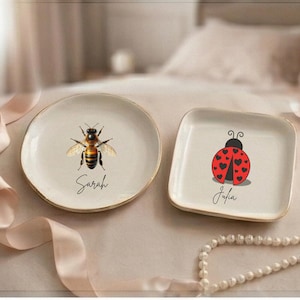 May include: Two decorative ceramic plates. One round plate features a bee illustration with the name "Sarah". The other is square and has a ladybug design with the name "Julia". Both plates have a gold rim.