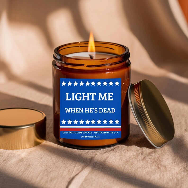 May include: A lit amber glass jar candle with a gold lid. The candle has a blue and white label that reads "LIGHT ME WHEN HE'S DEAD". The candle is made with natural soy wax and assembled in the USA.