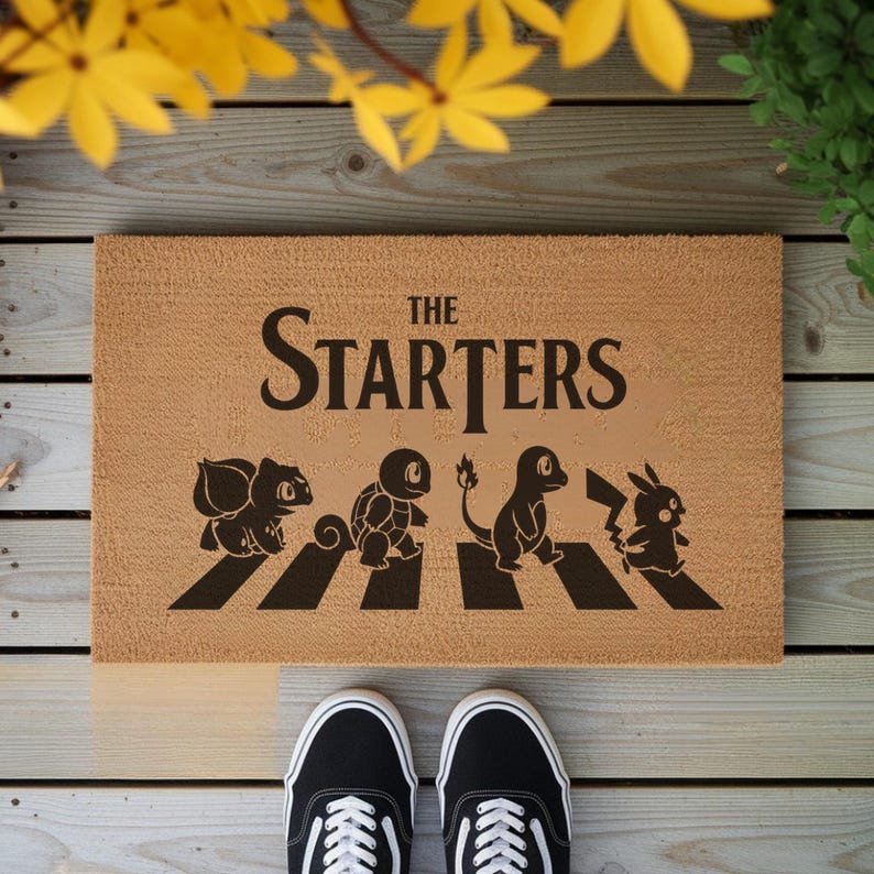 May include: A rectangular tan coir doormat featuring the black text THE STARTERS above a black graphic of four creatures walking across a crosswalk. The design includes silhouettes of Bulbasaur, Squirtle, Charmander, and Pikachu in a line.