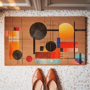 May include: A rectangular doormat with an abstract geometric design in shades of orange, red, black, blue, and yellow. The mat is made of a natural fiber material. Brown leather shoes are at the bottom of the image.