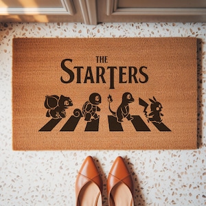May include: A rectangular tan coir doormat featuring the black text THE STARTERS above a graphic of four creatures walking across a crosswalk. The design includes silhouettes of Bulbasaur, Squirtle, Charmander, and Pikachu in a line.
