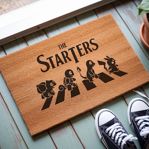 May include: A rectangular tan coir doormat featuring the black text THE STARTERS above a graphic of four cartoon creatures walking across black stripes. The design depicts Bulbasaur, Squirtle, Charmander, and Pikachu in a line formation.