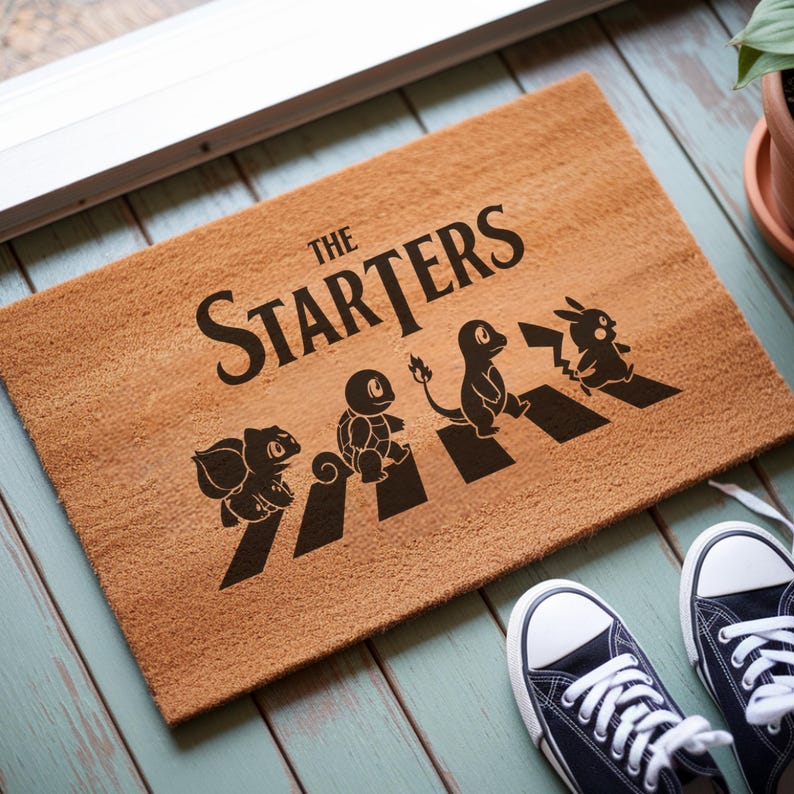 May include: A rectangular tan coir doormat featuring the black text THE STARTERS above a graphic of four cartoon creatures walking across black stripes. The design depicts Bulbasaur, Squirtle, Charmander, and Pikachu in a line formation.