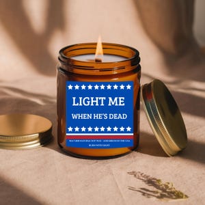 May include: A lit amber glass candle with a gold lid. The candle has a blue label with white text that reads "LIGHT ME WHEN HE'S DEAD." The candle is made with natural soy wax and assembled in the USA.