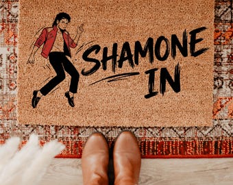 May include: A rectangular tan coir doormat featuring a black graphic illustration of a person in a red jacket and black pants performing a dance move, with the black text SHAMONE IN printed in a bold, brush-stroke style font on the right side.