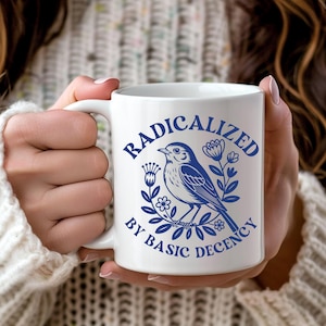 May include: White ceramic mug with a blue illustration of a bird and floral elements, with the text "RADICALIZED BY BASIC DECENCY" in a matching blue font. The mug has a white handle and is held by a person.