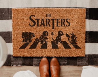 May include: A rectangular tan coir doormat featuring the black text THE STARTERS above a graphic of four creatures walking across black stripes. The design depicts Bulbasaur, Squirtle, Charmander, and Pikachu in a line, mimicking a famous album cover.